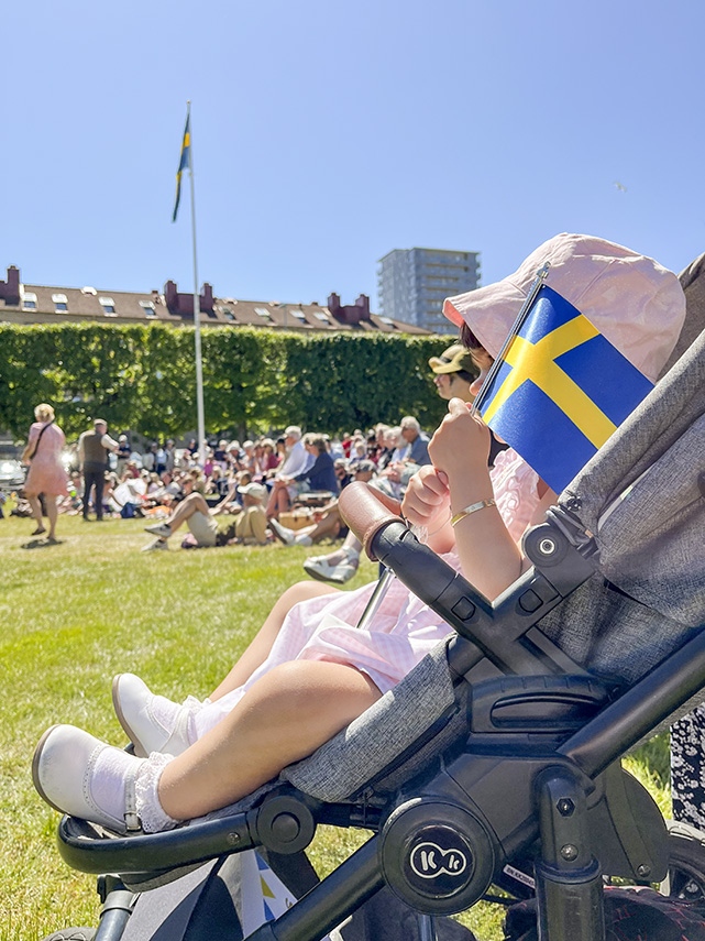 Liten flicka sitter i barnvagn och håller en liten svensk flagga i handen. 